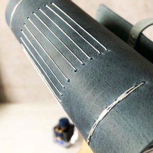May include: Close-up of a rolled, dark teal leather journal with white stitching. The stitching creates a series of parallel lines across the leather. A gray leather strap is visible on the right side. The journal appears to be handmade.
