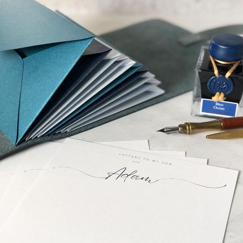 May include: A stationery set featuring a teal leather-bound envelope holder with multiple teal and navy envelopes. A bottle of blue ink and a calligraphy pen are also present. The stationery includes a letter addressed to "Adam".