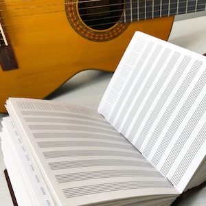 May include: A white music notebook with lined pages for writing music notes. The notebook is open and lying on a white surface next to a yellow acoustic guitar.