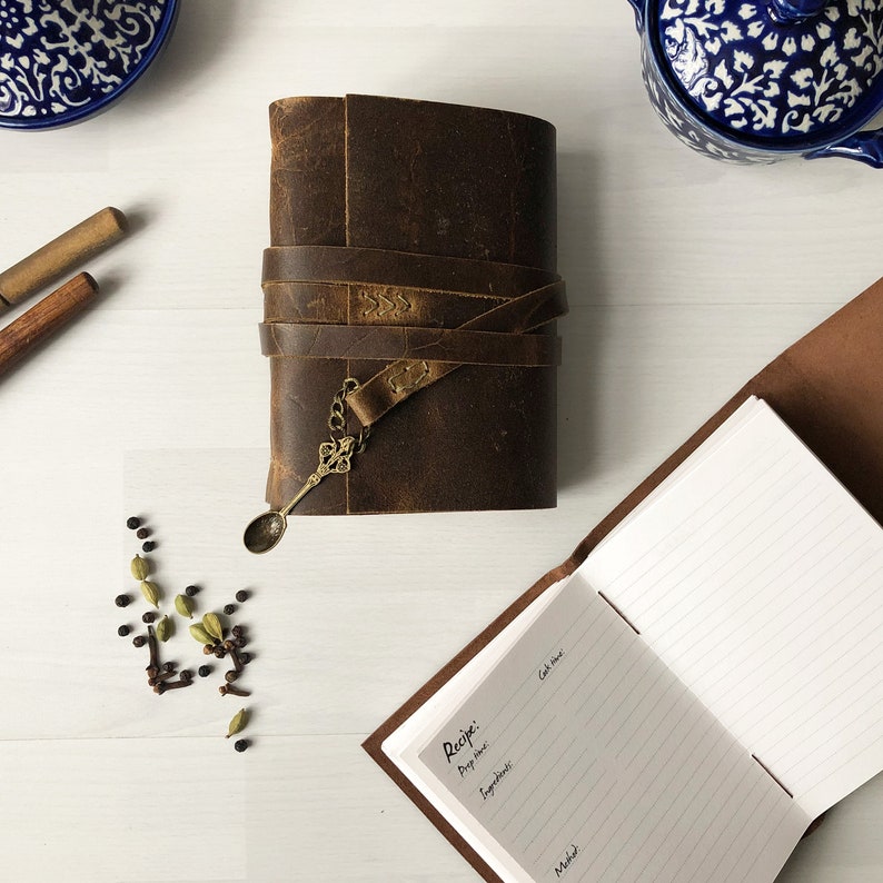 May include: A brown leather journal with a metal clasp and a small spoon charm. The journal is closed and has a rustic look. It is sitting on a white surface with a small pile of spices next to it.