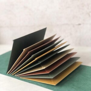 May include: An open, multi-colored photo album with pages fanned out. The pages are various shades of brown, gold, and gray, with a black cover. The album rests on a green surface, with a white brick wall in the background.