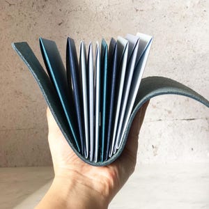 May include: A teal leather journal with a curved spine, held open to display the pages. The pages are a mix of white, teal, and navy blue. The journal's cover is a darker teal color, and the pages are fanned out.