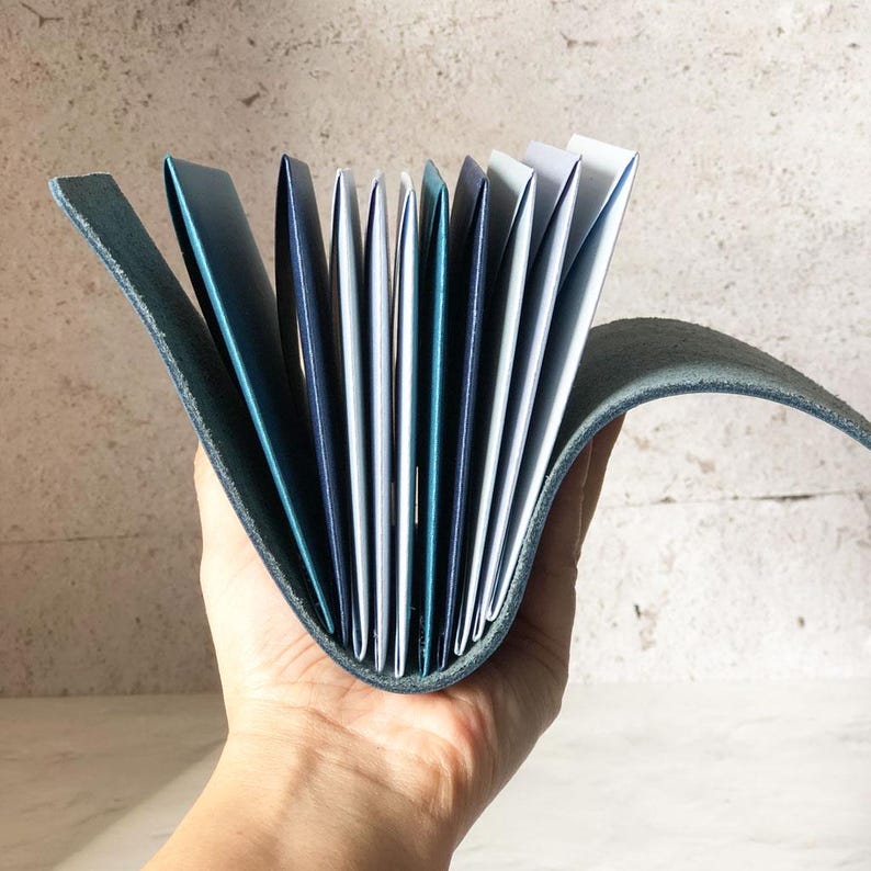 May include: A teal leather journal with a curved spine, held open to display the pages. The pages are a mix of white, teal, and navy blue. The journal's cover is a darker teal color, and the pages are fanned out.