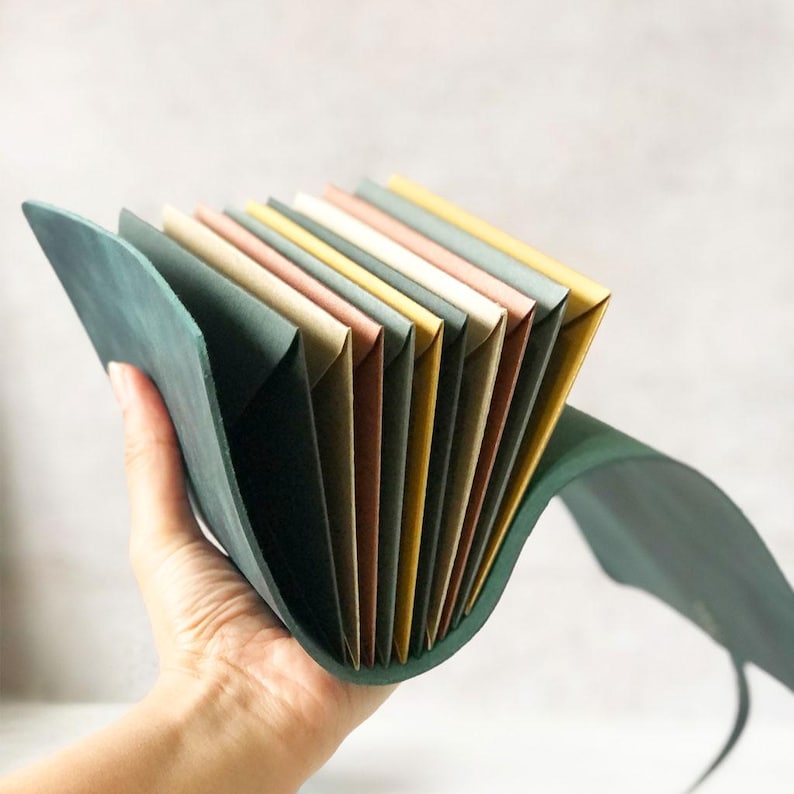 May include: A teal leather accordion file folder with multiple colored paper inserts. The file is held open, displaying the various pastel shades of the paper. The leather has a smooth texture.