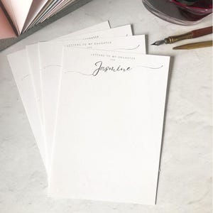 May include: A stack of white stationery sheets featuring the printed text "LETTERS TO MY DAUGHTER" and the name "Jasmine" in a black cursive script. The paper is arranged in a fan shape, accompanied by a fountain pen and an inkwell nearby.
