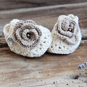 May include: A pair of crocheted baby booties with a flower design. The booties are cream colored with brown flower accents.