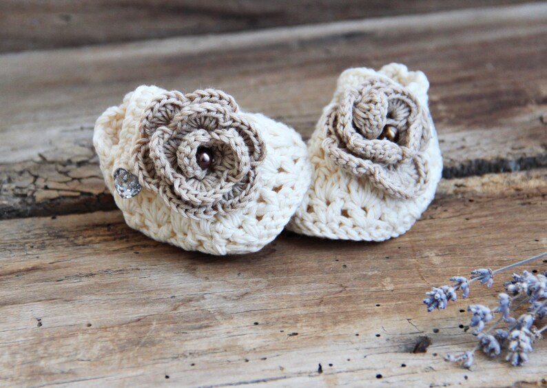 May include: A pair of crocheted baby booties with a flower design. The booties are cream colored with brown flower accents.