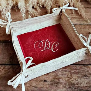 Pode incluir: Uma cesta de pão de linho branco com um forro de feltro vermelho e uma guarnição de renda branca. O forro tem um desenho bordado branco com as iniciais "DC".