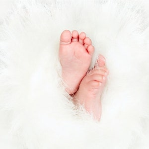 May include: A close-up of two tiny baby feet, nestled in a soft white fluffy blanket. The feet are pink and smooth, with tiny toes.