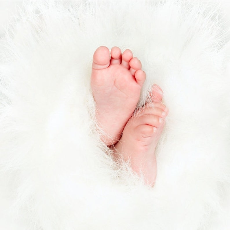 May include: A close-up of two tiny baby feet, nestled in a soft white fluffy blanket. The feet are pink and smooth, with tiny toes.