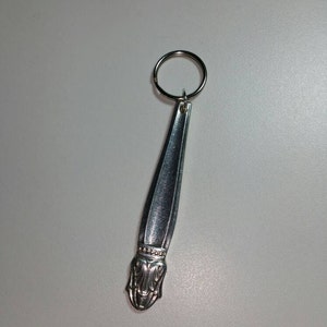 May include: A silver spoon keychain with a decorative design at the end. The spoon is attached to a silver key ring.