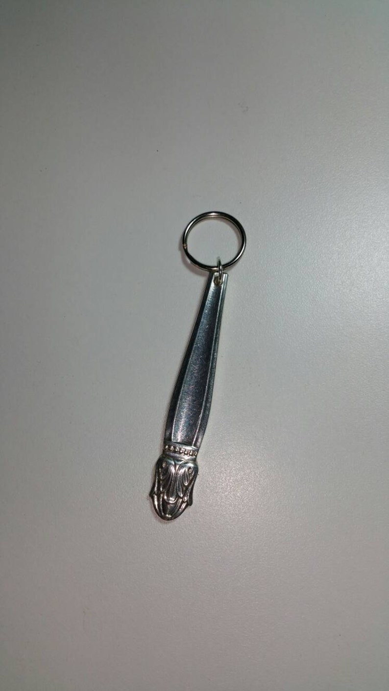 May include: A silver spoon keychain with a decorative design at the end. The spoon is attached to a silver key ring.