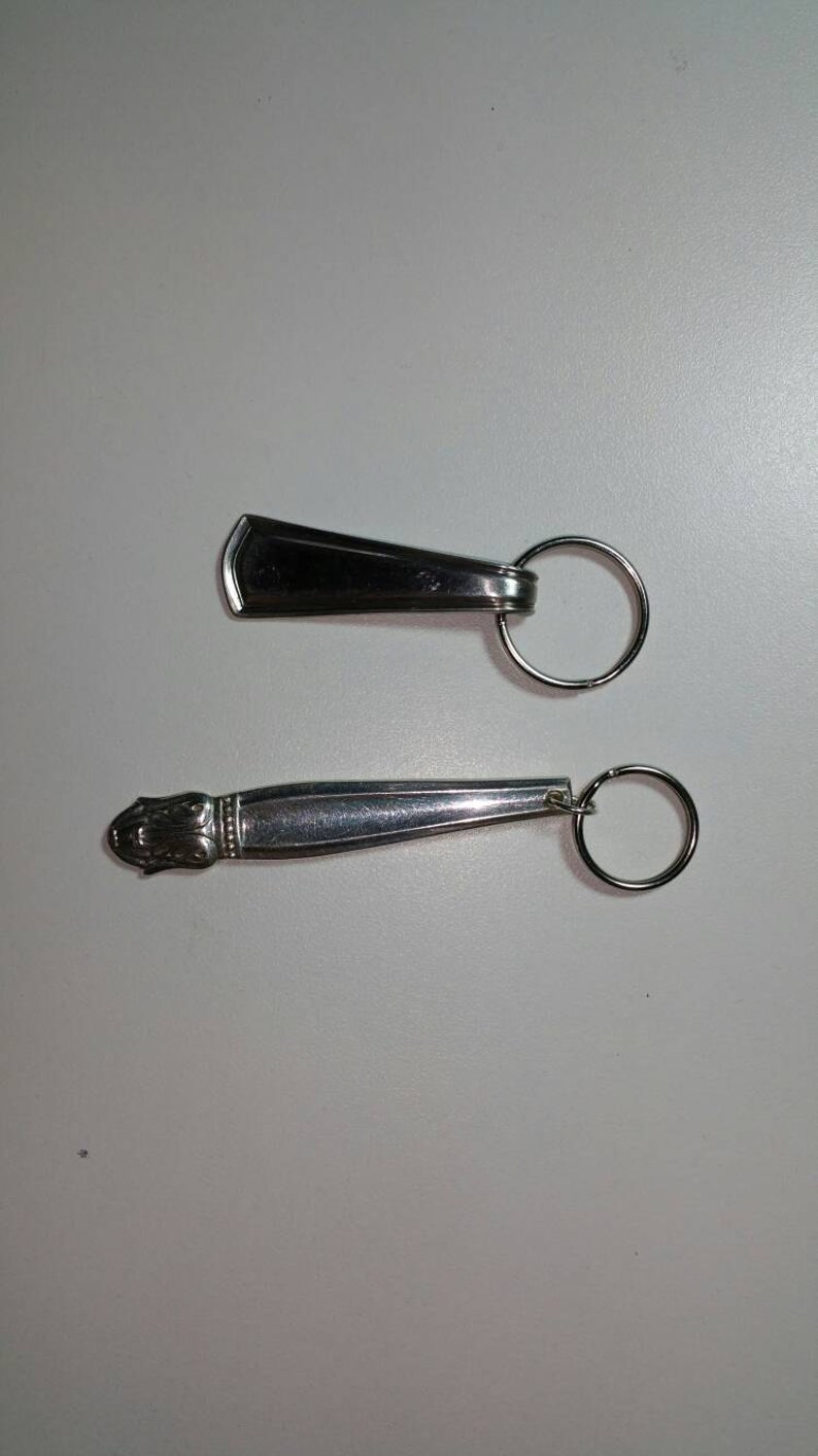 May include: Two silver keychains. The top keychain is a simple silver rectangle with a ring. The bottom keychain is a silver spoon with a decorative handle and a ring.