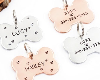 May include: Four personalized dog bone-shaped tags with engraved names and phone numbers. The tags are silver and copper in color. The names are Lucy, Harley, Abby, and Dori.