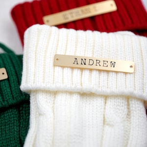 May include: Close-up of three knitted Christmas stockings in red, white, and green. Each stocking has a gold-coloured name tag. The white stocking displays the name "ANDREW". The red stocking has the name "ETHAN I".