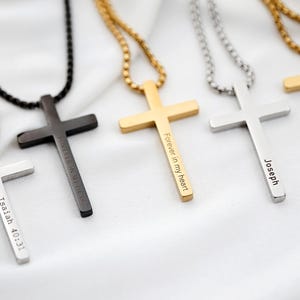 May include: Several cross pendants on chains in silver, black, and gold. Each cross has engraved text, such as "Isaiah 40:31", "Forever in my heart", "Joseph", and "Jeremiah 29:11". The necklaces are displayed on a white surface.