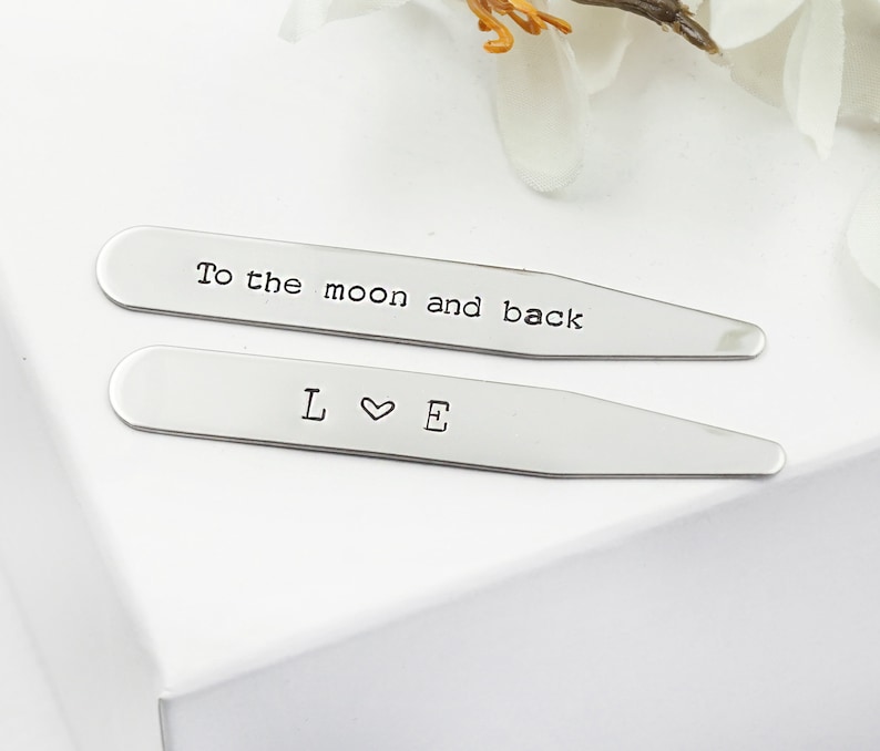 Op de afbeelding: Twee zilveren kraagstangen met gegraveerde tekst. De bovenste kraagstang luidt "To the moon and back". De onderste kraagstang luidt "L &hearts; E".