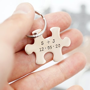 May include: A gold-coloured puzzle piece keyring with the initials "S + J" and the date "12.05.20" engraved. The keyring is attached to a silver metal ring.