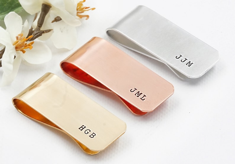 May include: Three personalized money clips in gold, rose gold, and silver. The clips are engraved with the initials "HGB", "JMI", and "JJM".