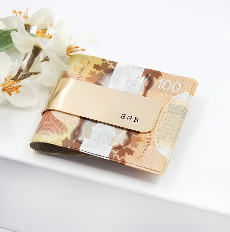May include: Gold money clip with the initials "HGB" engraved on it. The clip is holding a stack of Canadian $100 bills.