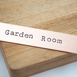 Peut inclure: Une plaque rectangulaire en métal couleur cuivre avec les mots "Garden Room" en noir, police sans-serif. La plaque a un trou à chaque extrémité. La plaque est posée sur une surface en bois.