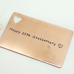 May include: A copper-coloured card with the text "Happy 25th Anniversary" and a small heart engraved on it. A silver necklace with a heart pendant engraved with the letters "SVR" is also pictured.