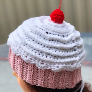 May include: A white and pink crocheted cupcake hat with a red cherry on top.