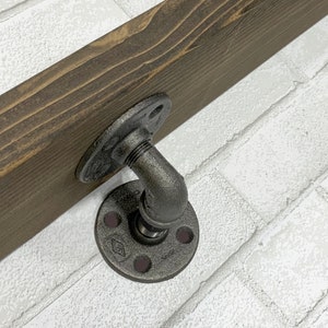 May include: A black metal pipe fitting with a flange on each end, attached to a dark brown wooden shelf. The shelf is mounted to a white brick wall.