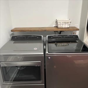 May include: A silver Whirlpool washing machine and tumble dryer set with a wooden shelf above. A wire basket with a white cloth liner sits on the shelf. The appliances are in a utility room.