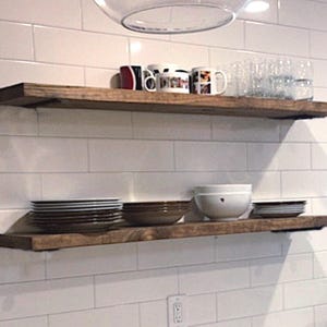 May include: Two rustic, wooden floating shelves on a white tiled wall. The top shelf holds mugs and glasses, the bottom shelf displays plates and bowls. The shelves are stained a warm brown colour.