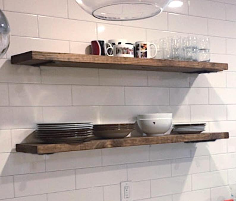 May include: Two rustic, wooden floating shelves on a white tiled wall. The top shelf holds mugs and glasses, the bottom shelf displays plates and bowls. The shelves are stained a warm brown colour.