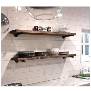 May include: Two horizontal wooden shelves mounted with black metal industrial pipe brackets. The top shelf holds various mugs and clear glasses. The bottom shelf holds stacks of plates, bowls, and a white ceramic bowl against a white tiled wall.