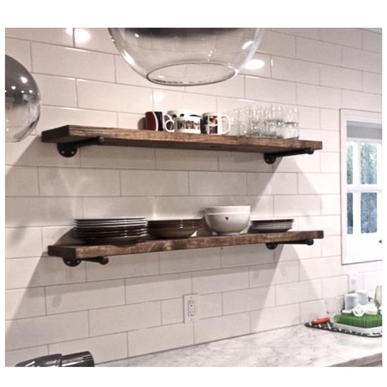 May include: Two horizontal wooden shelves mounted with black metal industrial pipe brackets. The top shelf holds various mugs and clear glasses. The bottom shelf holds stacks of plates, bowls, and a white ceramic bowl against a white tiled wall.