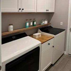May include: A utility room with white cupboards, black handles, and a wooden worktop. Two washing machines, one black and one white, are present. Shelves display jars and bottles with different contents.