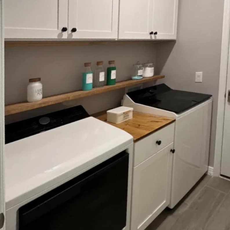 May include: A utility room with white cupboards, black handles, and a wooden worktop. Two washing machines, one black and one white, are present. Shelves display jars and bottles with different contents.