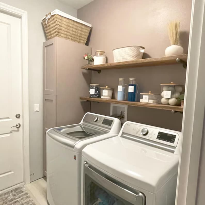 May include: A laundry room featuring two white top-loading washing machines and a dryer positioned beneath two wooden shelves. The shelves hold various glass storage jars with white labels, a white woven basket, and a white vase with dried grass.
