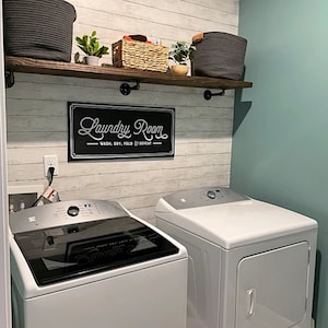 May include: A laundry room setup featuring a white top-load washer and dryer. Above the appliances, a dark wood shelf with black metal brackets holds two dark gray woven baskets and a small wicker basket. A black sign reads Laundry Room.