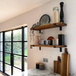 May include: Two wooden shelves with black metal brackets are mounted on a white wall. The top shelf holds a clock, a glass jar with grains, and a black pepper grinder. The bottom shelf displays a pink vase, mugs, and a black canister.