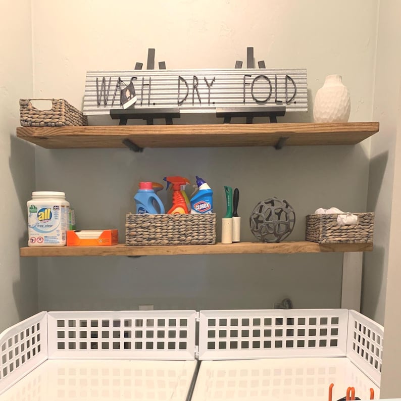 May include: A laundry room setup with two wooden shelves displaying cleaning products and decor. The top shelf has a sign saying "WASH. DRY. FOLD." and a white vase. The lower shelf holds detergent, spray bottles, and a decorative sphere, all arranged in a functional space.