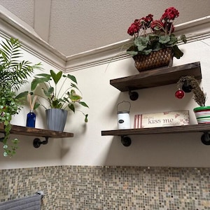 May include: Two dark wood floating shelves mounted with black metal pipe brackets. The shelves hold a blue glass bottle, a gray ribbed planter with a green plant, a brown woven basket with red flowers, a white lantern, and a sign reading Always kiss me goodnight.