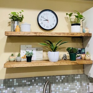 May include: Two wooden shelves hold potted plants, a black clock, and ornaments. The clock shows the time as 9:47. The shelves are mounted on a light-coloured wall above a tiled backsplash. The shelves are made of rustic wood.