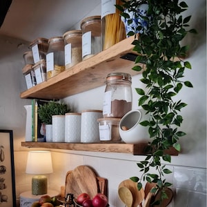 May include: A kitchen scene featuring two wooden shelves filled with various glass jars and white ceramic containers. The jars contain dry goods, and the ceramic containers have wooden lids. A trailing green plant hangs from the top shelf.