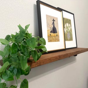 May include: A rustic wooden floating shelf holds two framed art prints and a trailing green artificial plant. The left print depicts a person in a dark dress and hat carrying a basket, while the right print shows white flowers in a vase.