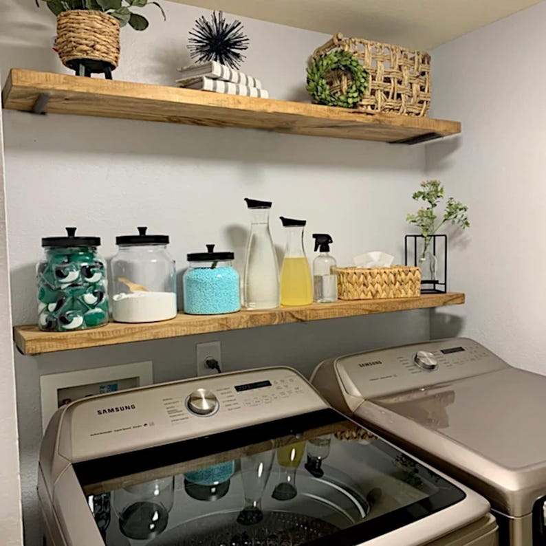May include: A well-organised laundry room featuring two wooden shelves stocked with cleaning essentials. The shelves are positioned above two silver washing machines. Jars of detergent, fabric softener, and spray bottles are visible, alongside decorative baskets and plants.