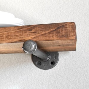 May include: A wall-mounted shelf featuring a thick, rectangular wooden board with a visible grain pattern, supported by a dark gray metal pipe bracket. The bracket includes a circular flange with the text 1/2 clearly visible on the metal surface.