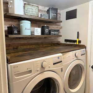 May include: A laundry room with a washing machine and dryer. Wooden shelves hold various containers labeled "Pods," "Laundry," and "Dryer Sheets." Decorative signs with text like "Wash-Dry-Fold" and "The Laundry is Looking at Me Dirty Again" are also present.