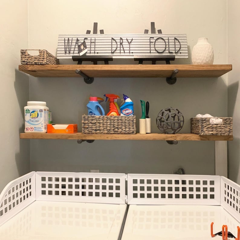 May include: Two wooden shelves with metal pipe brackets mounted on a wall. The top shelf holds a sign reading WASH. DRY. FOLD., a woven basket, and a white vase. The bottom shelf holds cleaning supplies, woven baskets, and a decorative sphere.