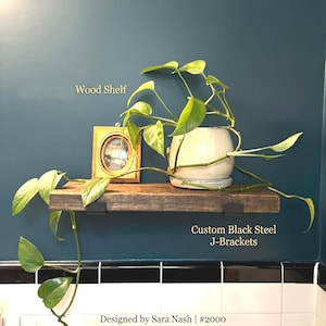 May include: A dark brown wooden shelf mounted with two custom black steel J-brackets. A trailing green pothos plant in a white ceramic pot and a small oval gold-toned picture frame sit on the shelf. Text reads Wood Shelf, Custom Black Steel J-Brackets, Designed by Sara Nash | #2000.