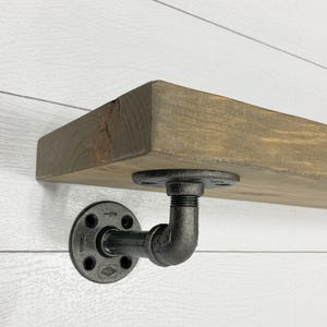 May include: A thick, rectangular wooden shelf with a dark brown stained finish, supported by a black industrial-style metal pipe bracket. The bracket consists of a circular wall flange and a threaded elbow fitting attached to the underside.