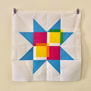 Op de afbeelding: Een vierkant stoffen quiltblok met een centrale achtpuntige ster in felblauw op een witte achtergrond. Het midden van de ster bevat vier kleinere vierkanten in magenta, geel, oranje en groen, gerangschikt in een rasterpatroon.
